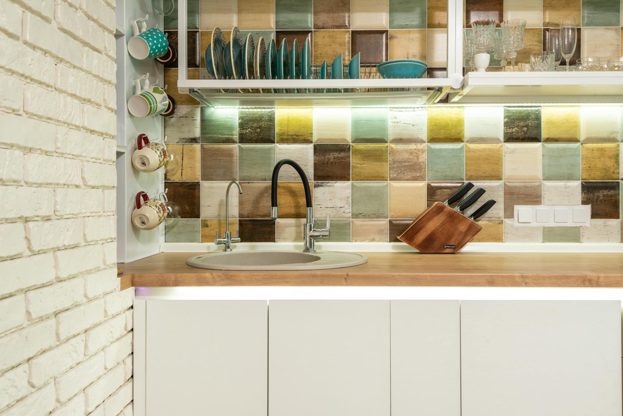 Interior of bright kitchen corner furnished with wooden counter and white cupboards decorated with various dishware and glasses against tiled wall